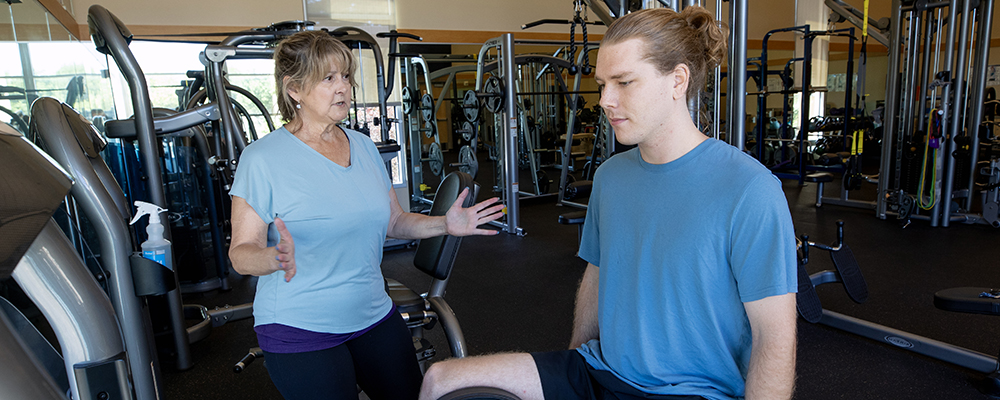An instructor in gym clothes speaks to a student seated on an adductor machine.