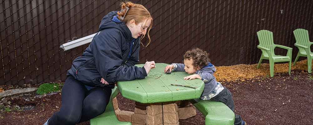 An adult caretaker smiles at a small child opposite her, playing with a toy car on the tabletop.