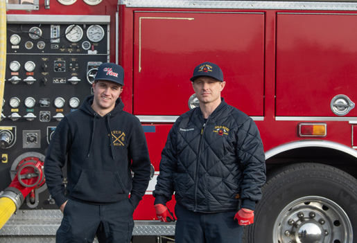 Two men wearing casual clothing with firefighting logos stand looking at the camera, with a red fire engine partially in view behind them.