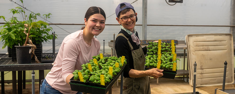 Two smiling students holding trays of basil plant starts pose in a large greenhouse.