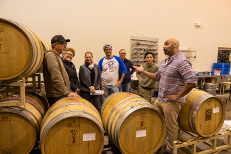 A group of wine studies students gather amongst oak wine barrels, listening to an expert lecture.