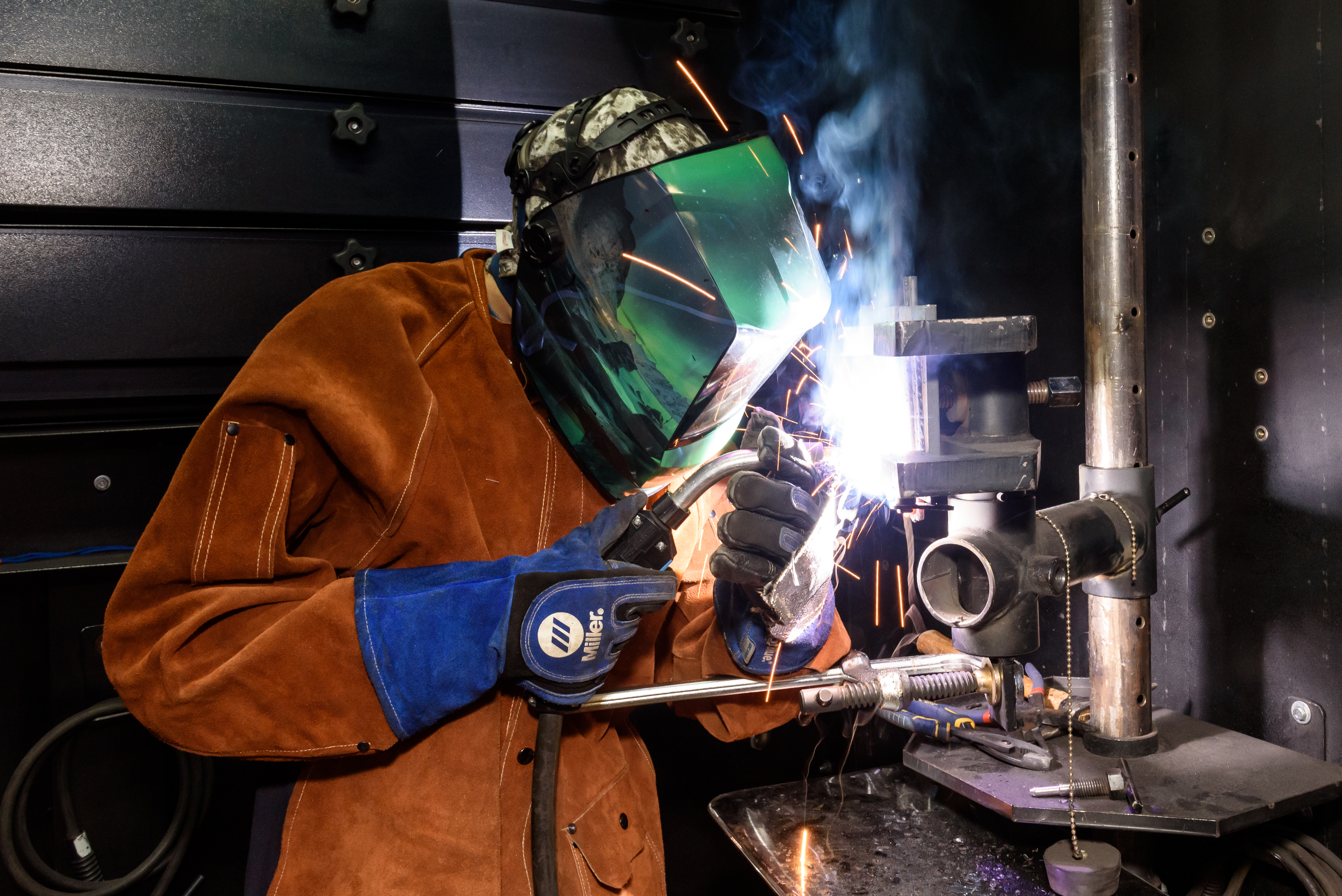 A student in welding helmet and heavy leather protective jacket and gloves focuses intently on using the welding torch in their hands.